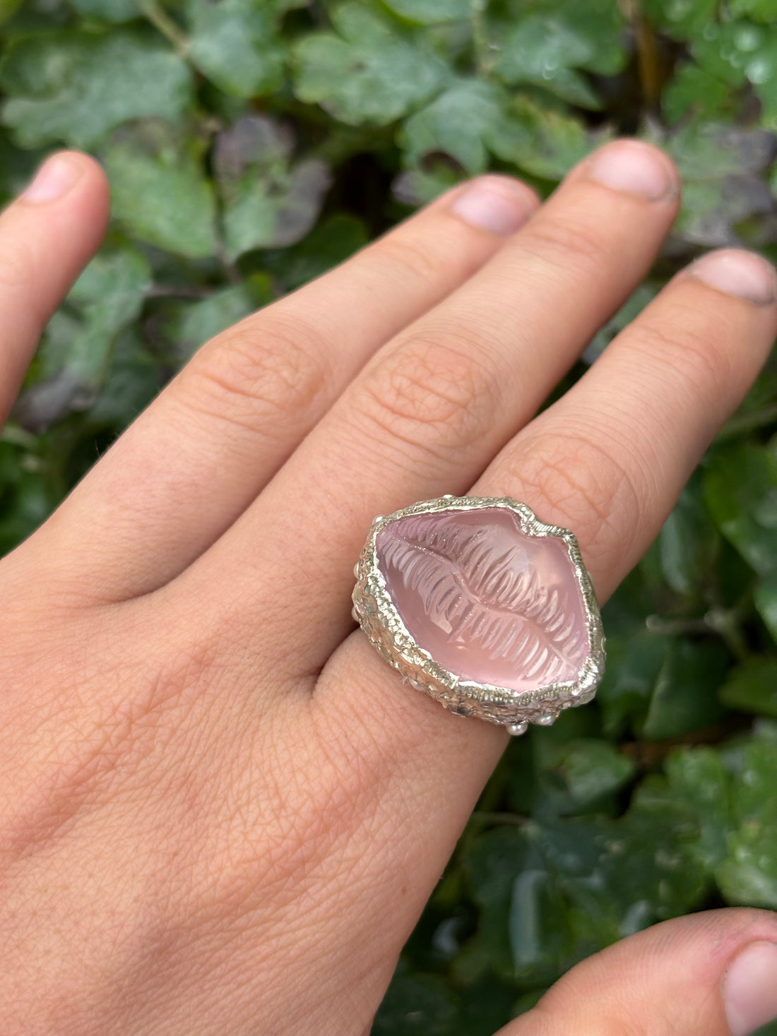 Hand wearing a ring with a pink stone against a green leafy background