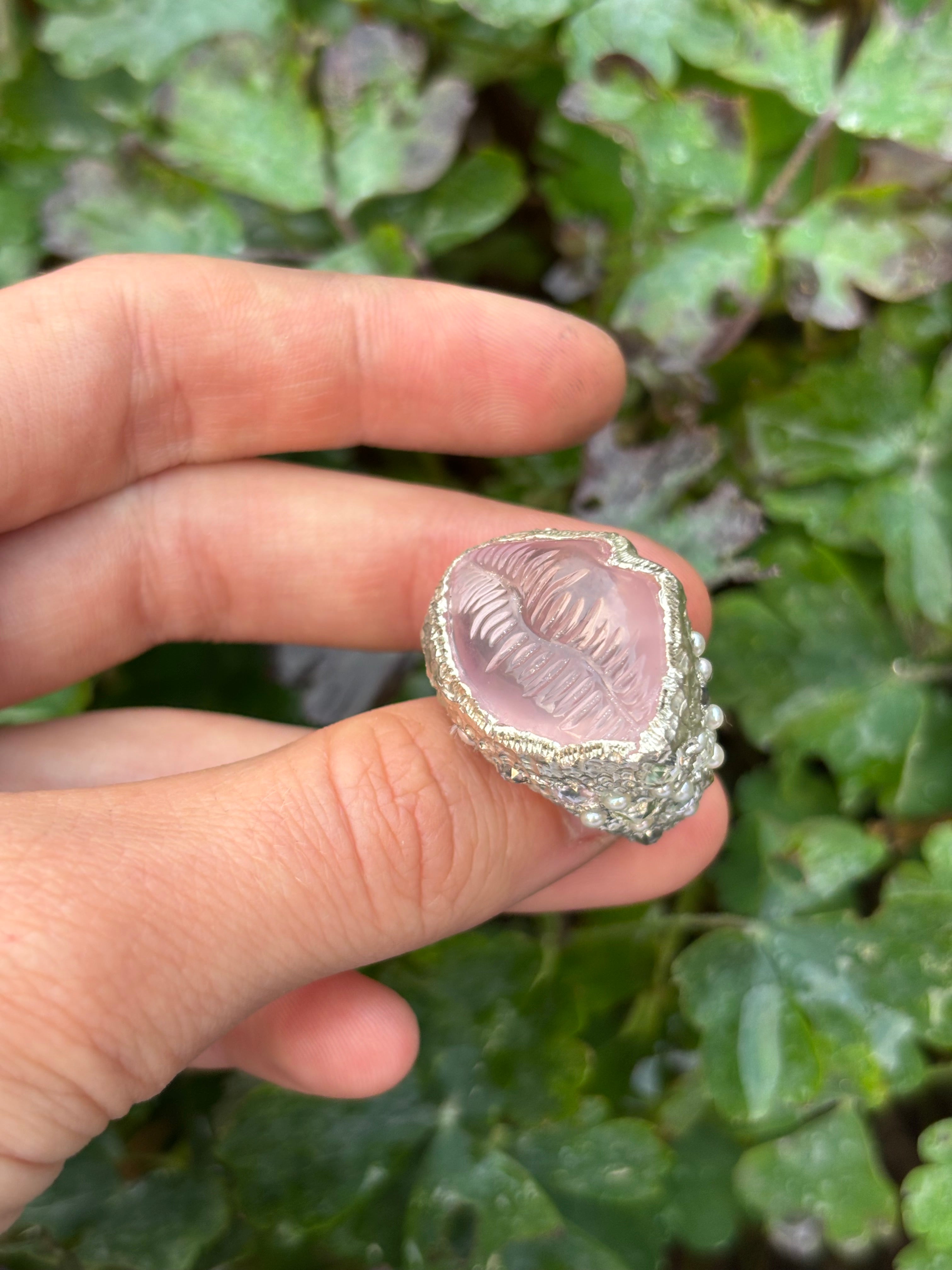 Hand holding a silver ring with a pink stone against a green leafy background