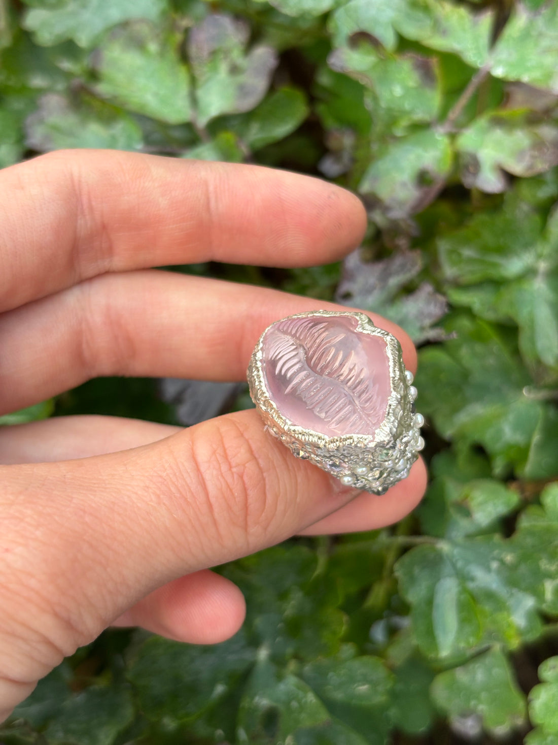 Hand holding a silver ring with a pink stone against a green leafy background