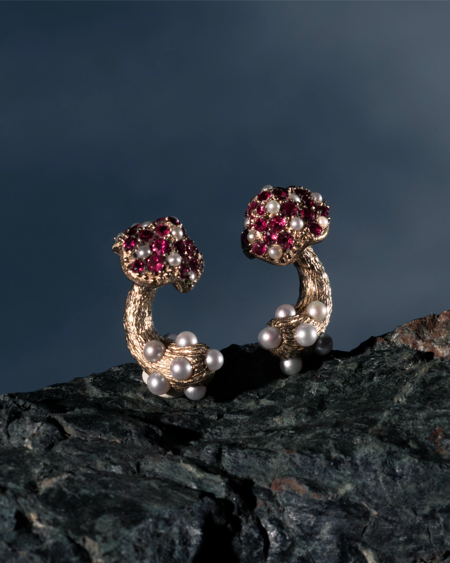 Pair of gold earrings with red gemstones and pearls on a dark rock surface